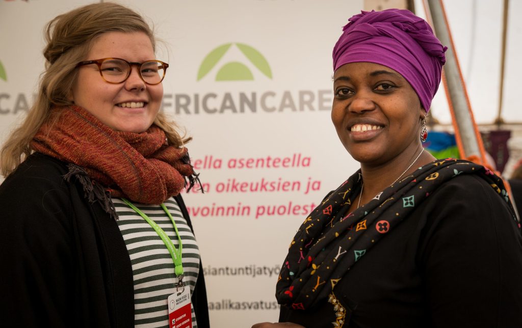 Kaksi naista seisovat vastakkain ja hymyilevät kameraan. Taustalla heidän välissään African care -järjestön roll-up.