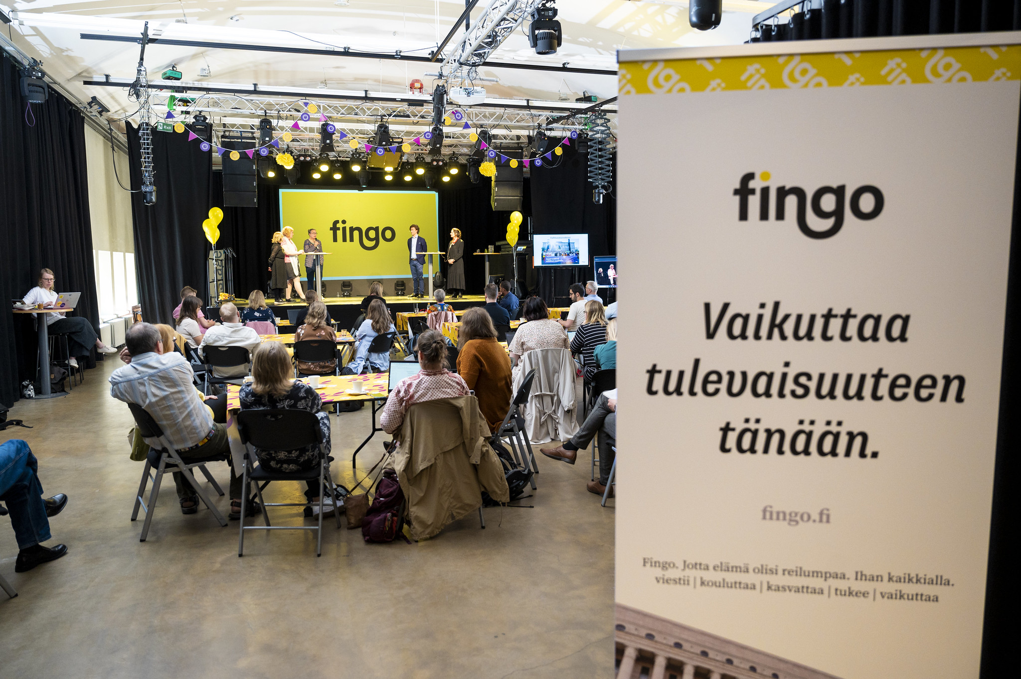 Fingon kevätkokousta kuvattuna yleisön takaa lavalle päin. Edustalla roll-up, jossa lukee Fingo, Vaikuttaa tulevaisuuteen tänään.